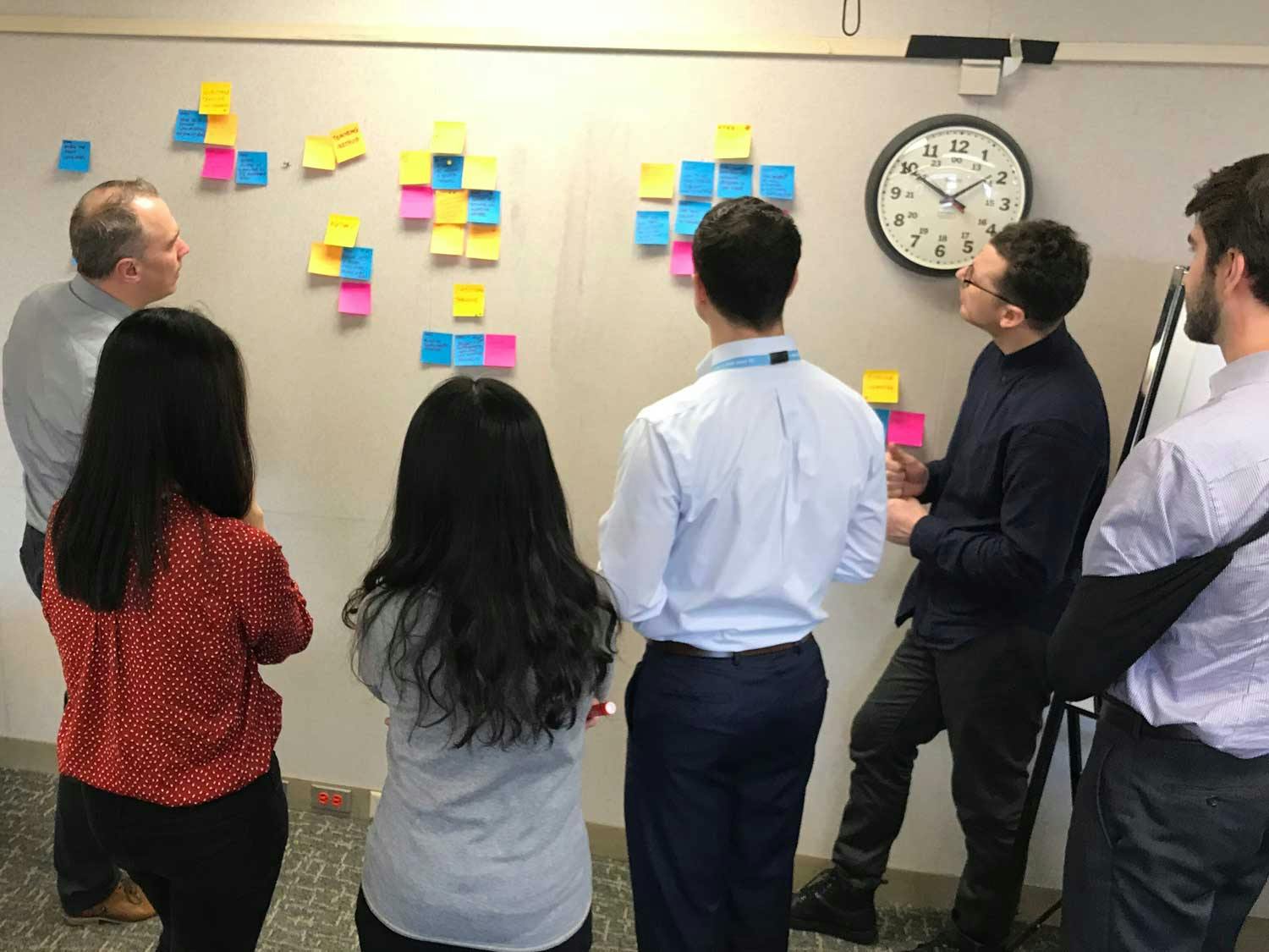 6 people gather around sticky notes on a white-board